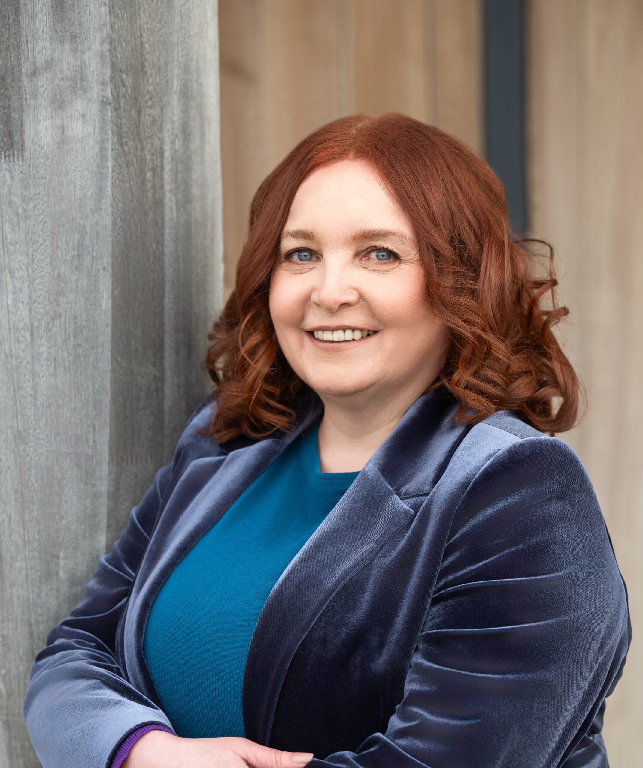 A person with curly red hair smiles, wearing a blue velvet blazer and teal top, leaning against a wooden surface.