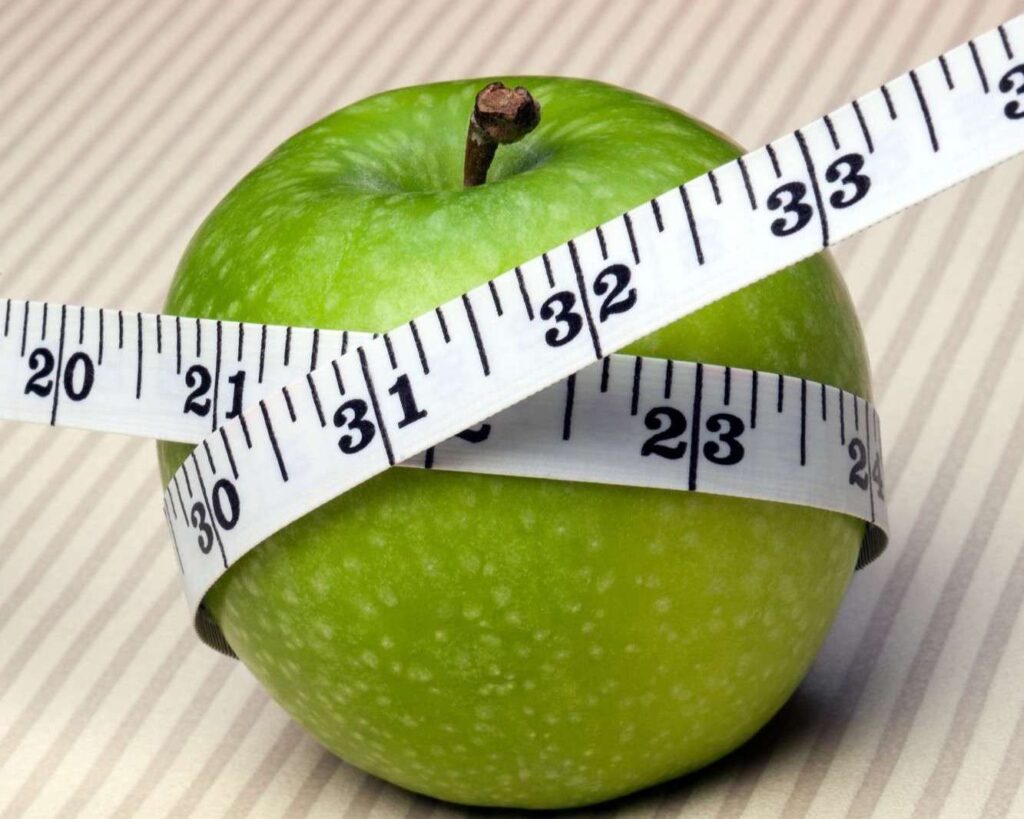 A green apple with a measuring tape wrapped around it, set on a surface with diagonal beige and white stripes.