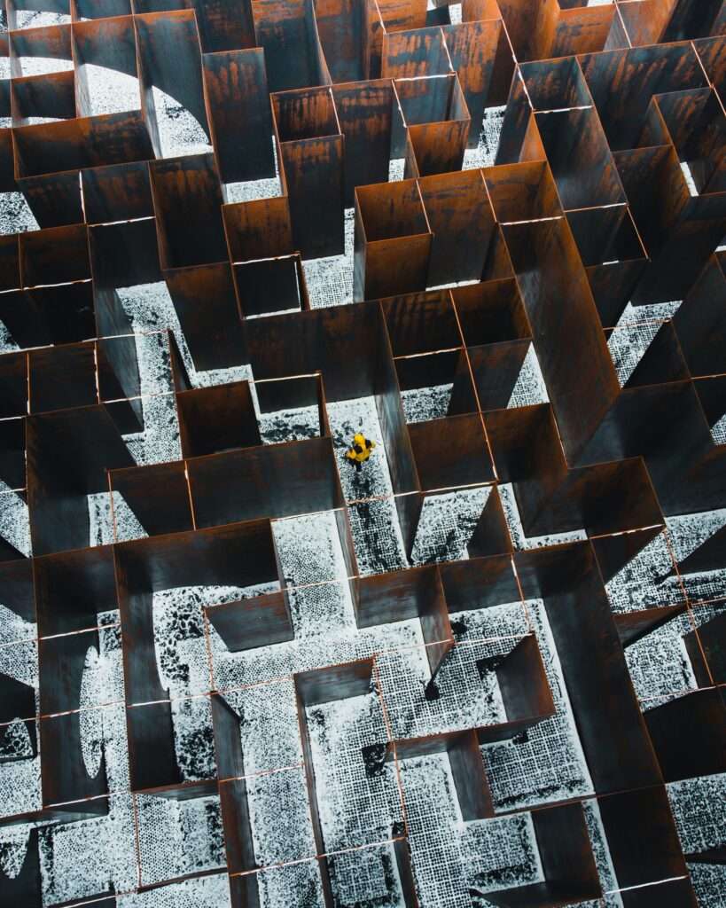 A person in a yellow jacket stands in the middle of a large, rust-coloured metal maze viewed from above.