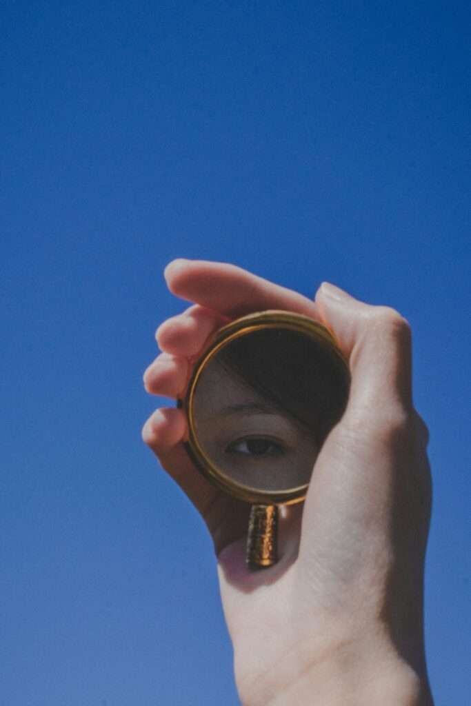 A hand holds a small round mirror reflecting one eye against a clear blue sky background.
