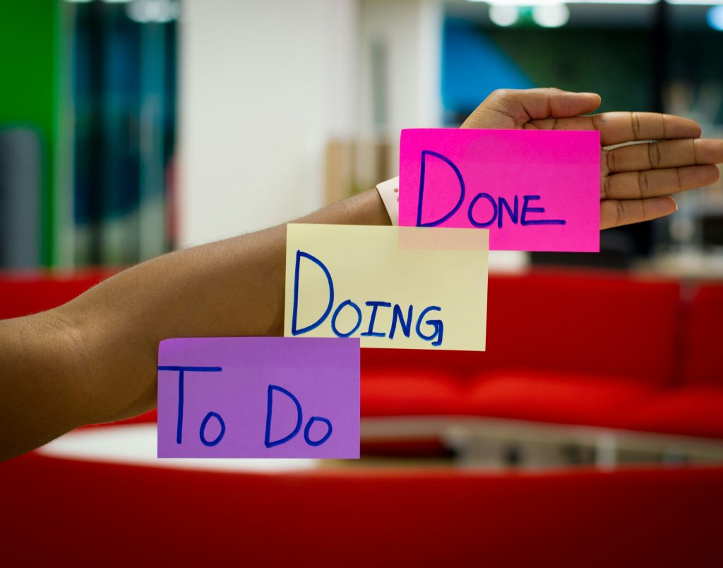 A hand holds three sticky notes labelled “To Do,” “Doing,” and “Done” in front of a blurred office background with red sofas.